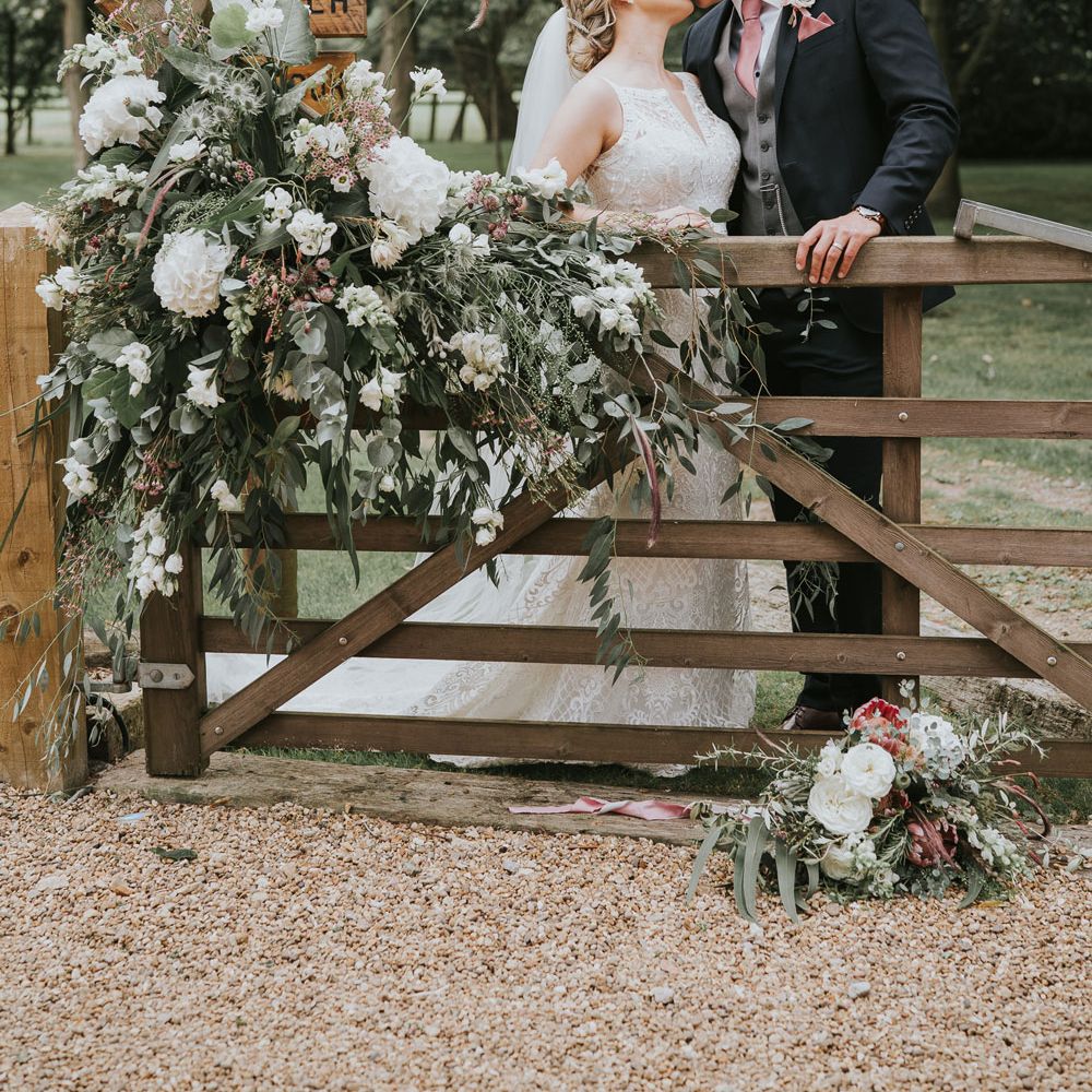 Bride in white lace Justin Alexander wedding dress kisses groom in dark three piece suit with grey waistcoat and pink tie behind wooden gate with white, pink and green floral drapery at Tythe Barn wedding with barn wedding flowers