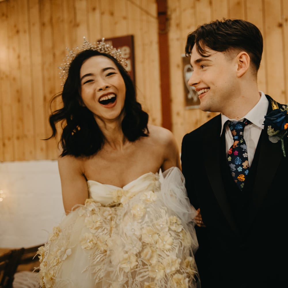 Bride in strapless wedding dress and bewjelled headpiece laughs with groom in blue suit, floral tie and floral buttonhole for rustic wedding
