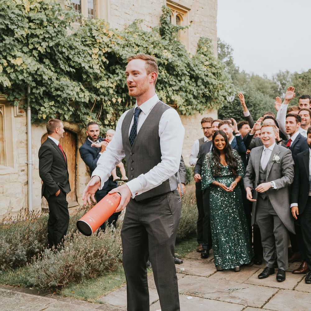 Groom in grey waistcoat and suit trousers gets ready to throw whisky bottle box to groomsmen, wedding guests and women in green sequin dress