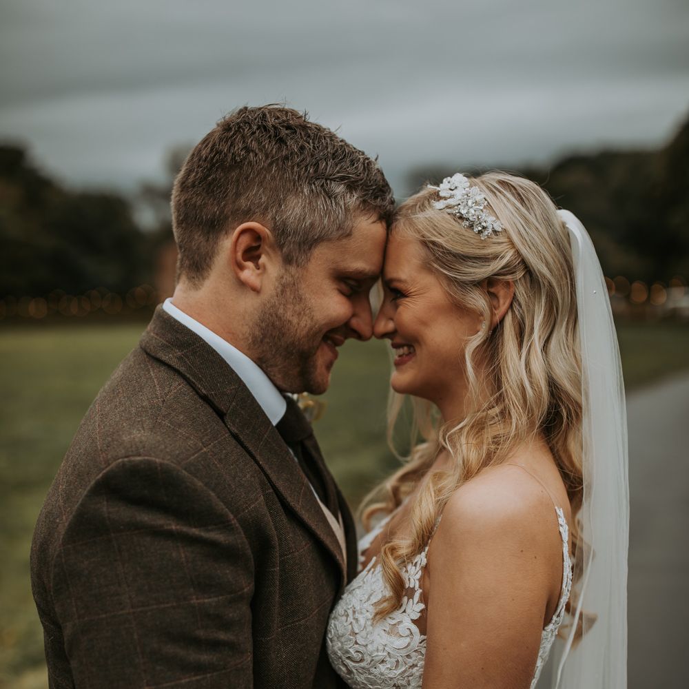 Bride in lace wedding dress and veils stand facing groom in brown check suit outside after barn wedding ceremony