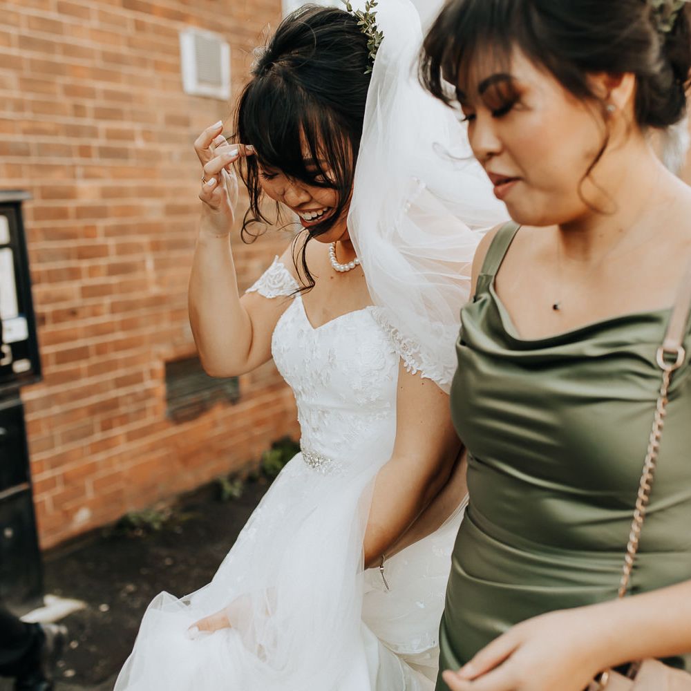 Bride in lace bardot wedding dress with pearl necklace and veil walks with bridesmaid in sage green satin bridesmaid dress towards wedding venue
