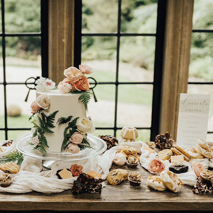 Dessert tables with biscuits, cakes and cookies, and a single tier buttercream wedding cake decorated with flowers and foliage 