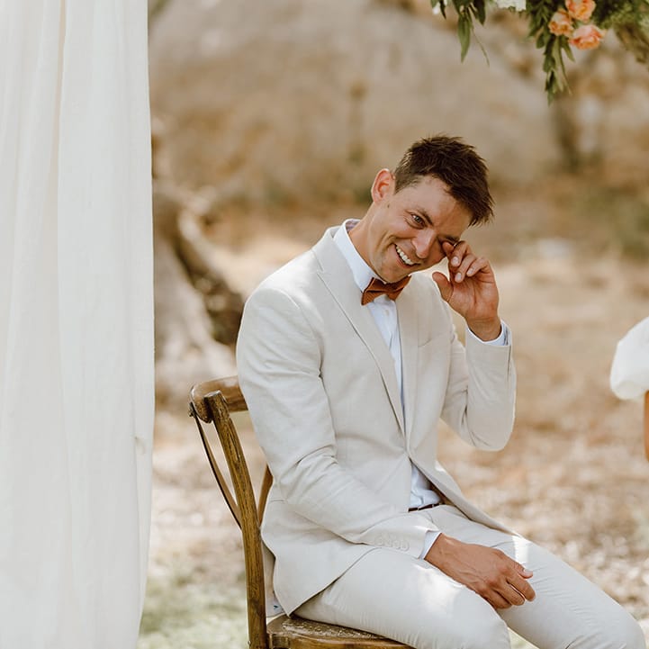 Bride and groom smiling and seated on wooden chairs in outdoors wedding