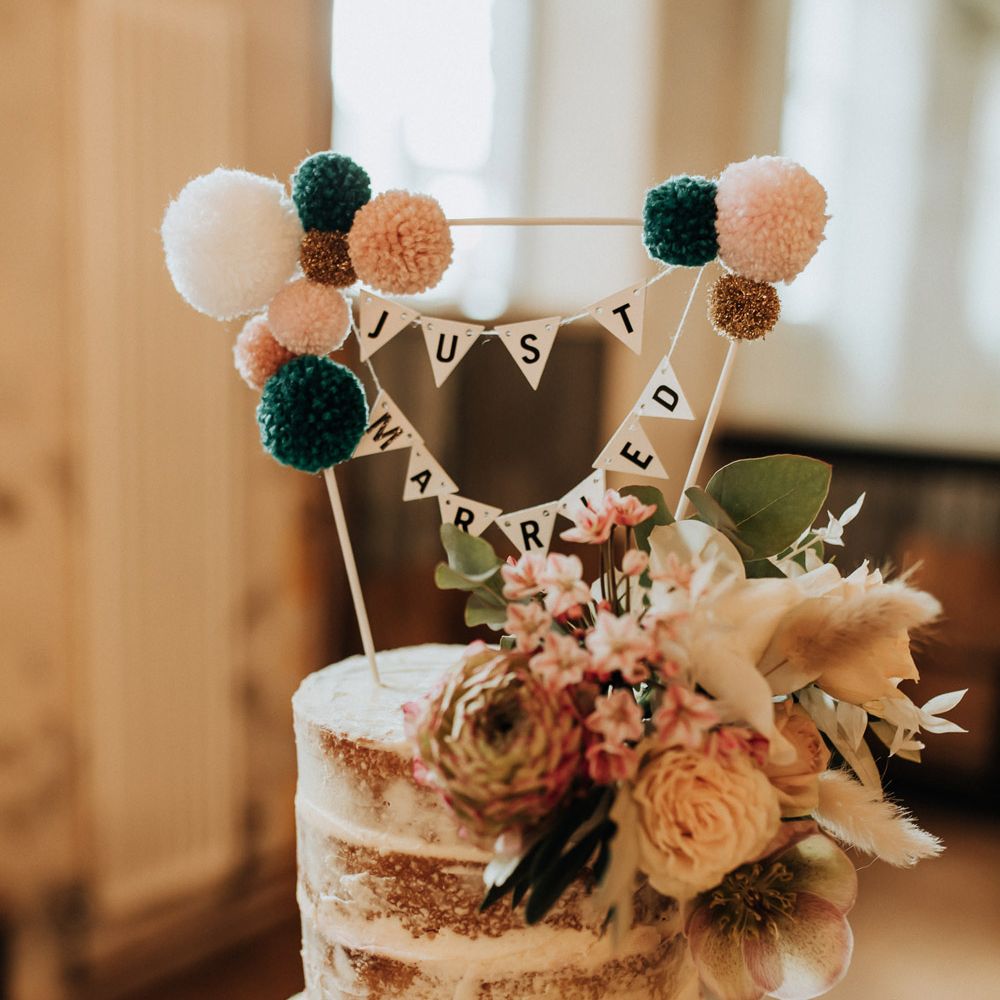 Rustic semi naked wedding cake with floral decor, bunting wedding topper reading 'Just Married' and pom poms