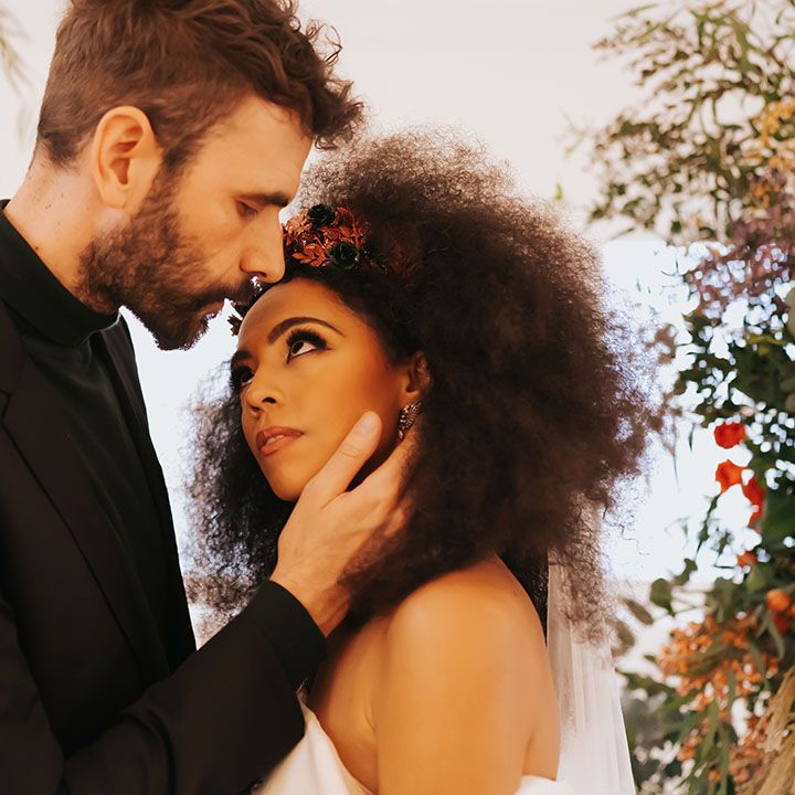 Groom in a black suit and top embracing his Black brides face with afro hair, off the shoulder wedding dress and winged eyeliner 