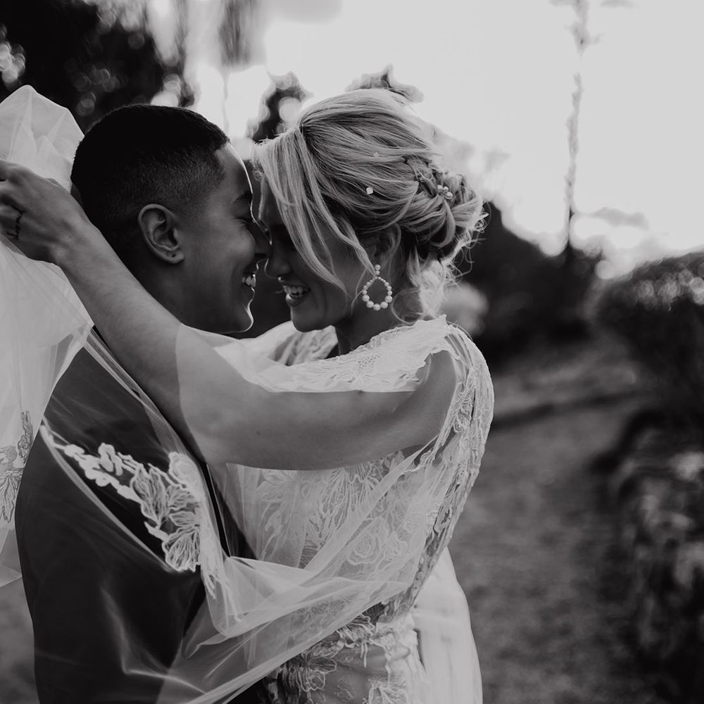 Bride in a lace wedding dress with pearl hoop earrings and pearl hair pins embracing her partner in a black tuxedo 