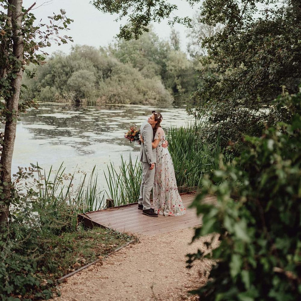 Bride in floral embroidered dress hugs groom in grey suit on jetty next to lake at wedding venue 