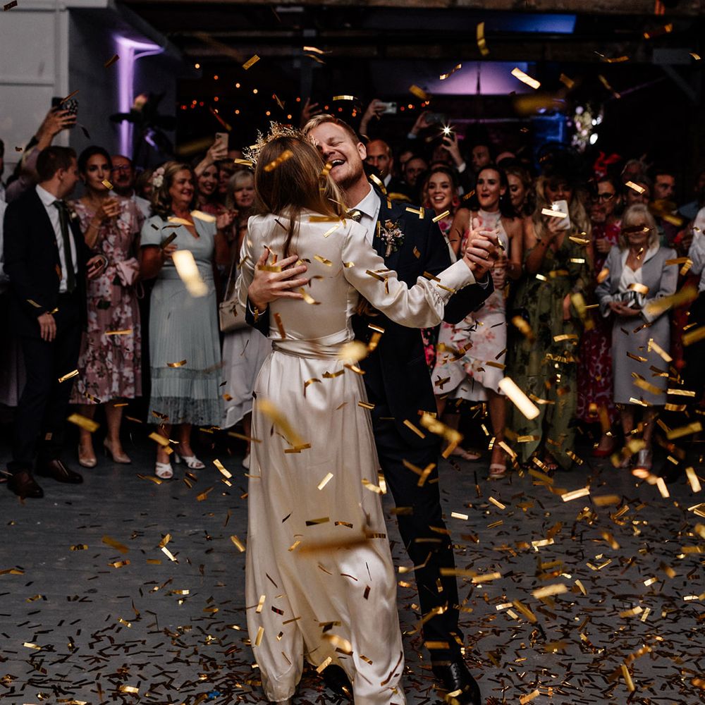 First dance with bride in a satin wrap wedding dress and gold crown and groom in a navy suit with gold confetti falling around them 