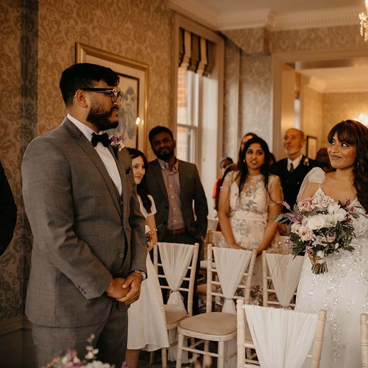 Bride in white traditional wedding dress (before mint green saree) and mixed neutral colour floral bouquet and groom is dark grey suit with bow tie 