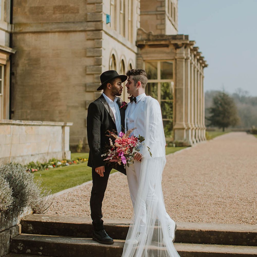 Groom in a two piece black suit and fedora hat standing on the steps at Prestwold Hall with his partner in a white three piece groom suit, white boots and sheer cape 