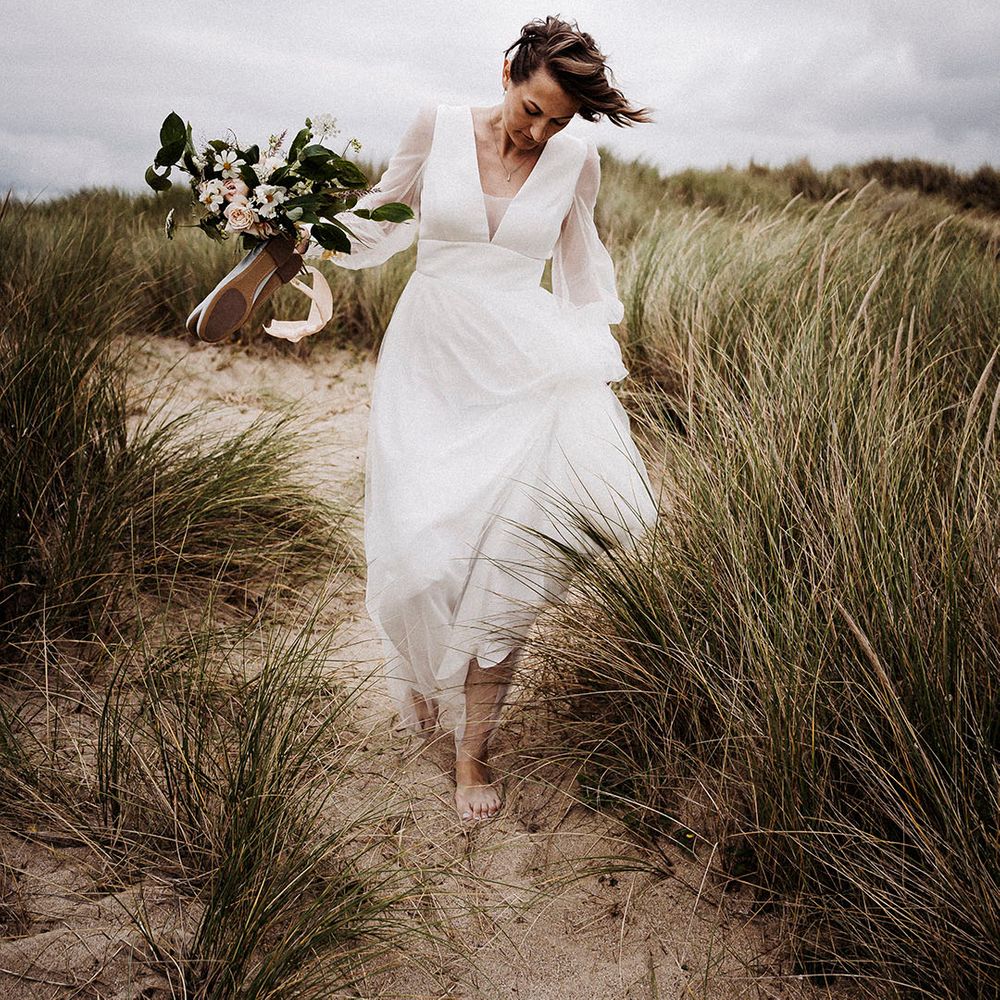 Bride holds her bouquet and wedding shoes as she walks in sand towards the beach in tulle Sassi Holford wedding dress