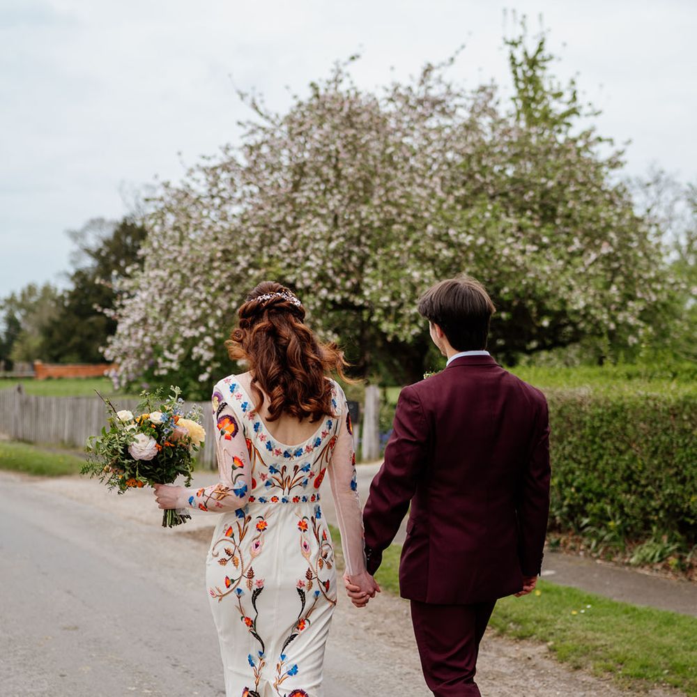 Groom in burgundy suit walks holding hands with bride in colourful wedding dress and bouquet 