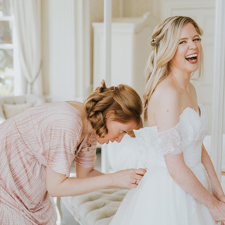 Bridesmaid buttons up the bride in her lace off the shoulder wedding dress with long blonde hair 