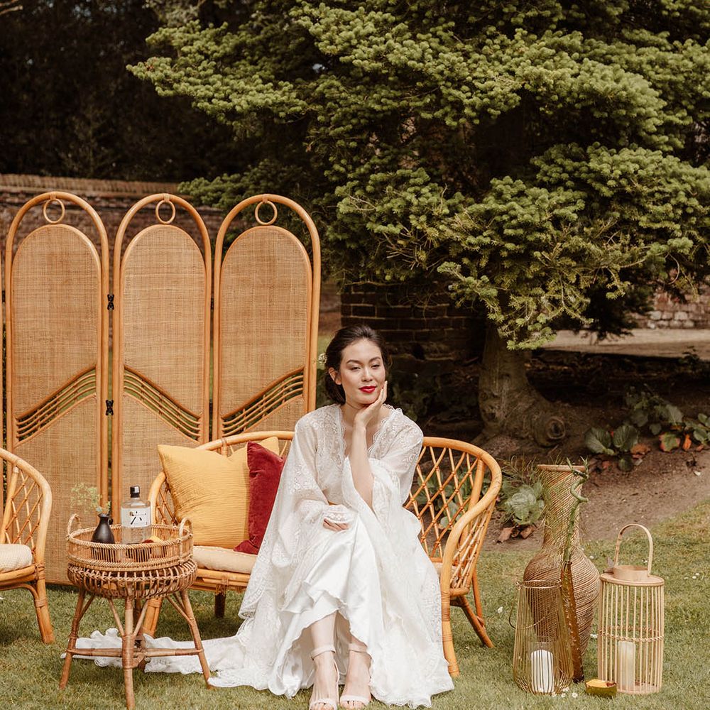 East Asian bride in a satin slip wedding dress with lace kimono style overlay sitting on a bamboo chair surrounded by a bamboo screen and table 