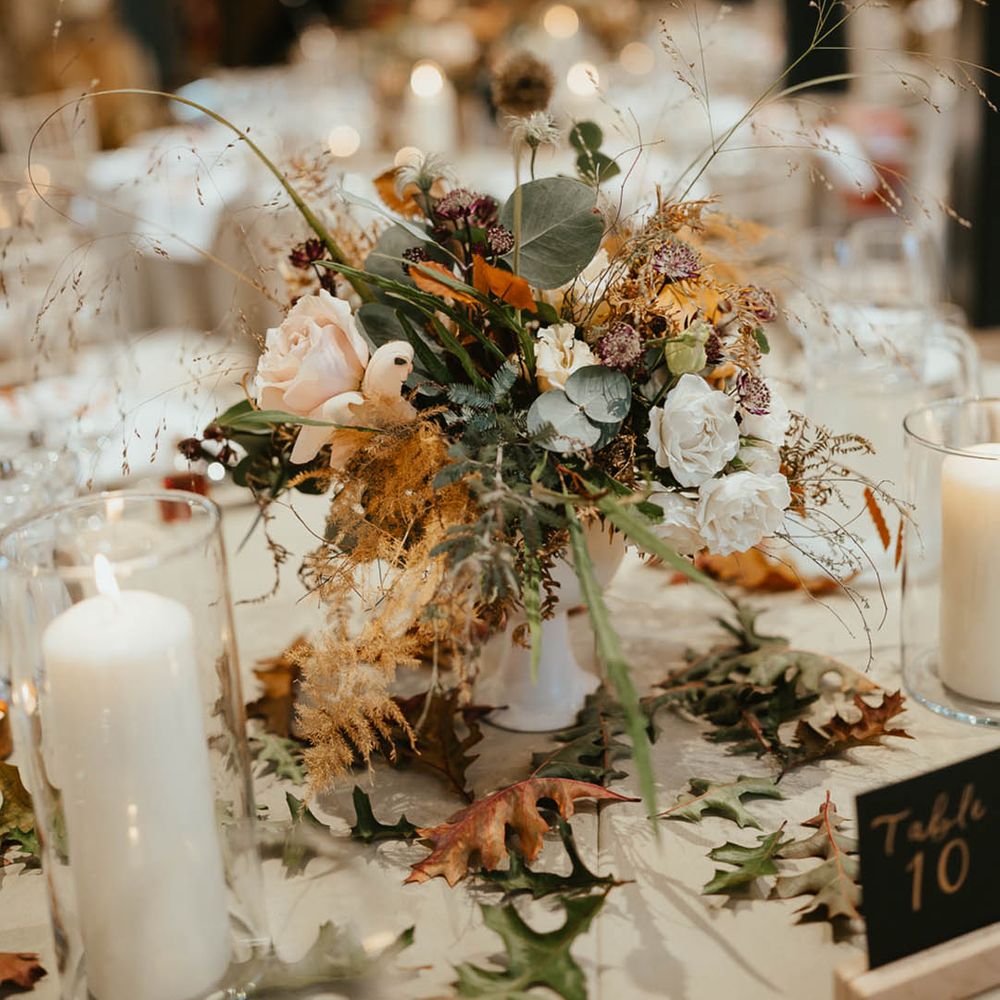 Wedding table centrepiece with autumnal leaves, white roses and dried grasses with white candles 