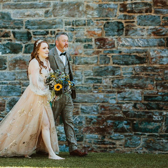 Bride wearing pink starry wedding dress walks with her father who wears three piece tartan suit with black bow tie and white shirt