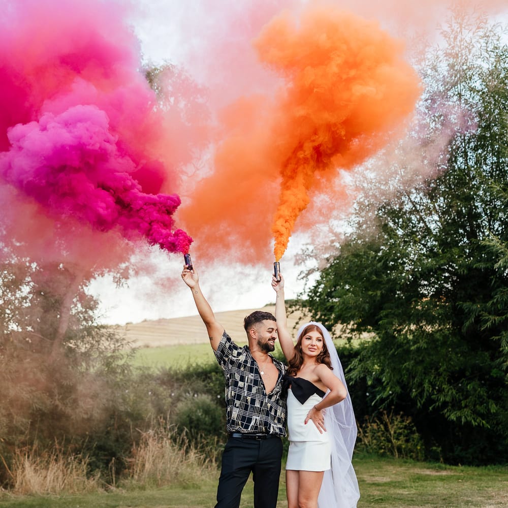 Bride & groom hold orange and pink smoke bombs outdoors for couples portraits 