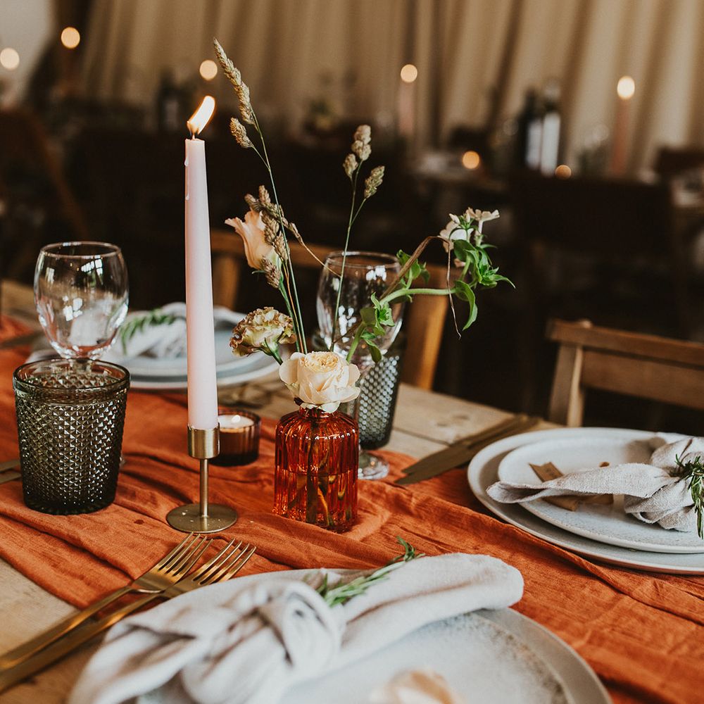 Flower stems in orange vase with candles, an orange table runner, and white plating 