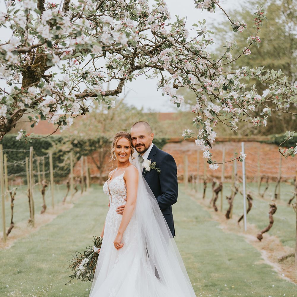 Bride with pearl drop earrings and floral lace wedding dress is embraced from behind by the groom 