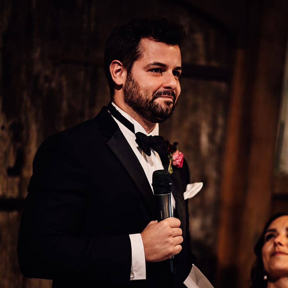 The groom in traditional black tie stands up with a microphone as he reads out his speech 