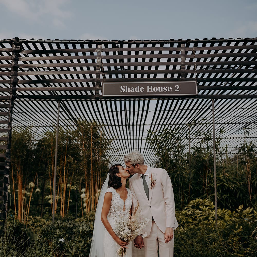 Bride and Groom kiss in front of greenhouse wearing a lace and tulle fishtail dress and a light pink suit