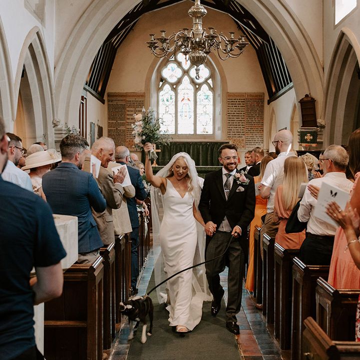The bride and groom walk back down the aisle as a married couple with their pet dog, Brains 