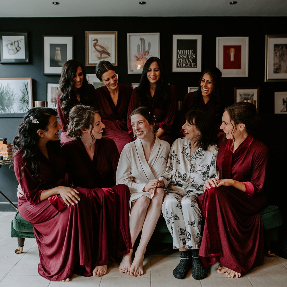 Bridesmaids in dark red satin robes, bride in cream robe with black stitching and mother of the bride in floral patterned silk pyjamas getting ready before wedding