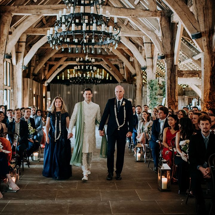 Groom walks down the aisle with his mother and father during barn wedding complete with festoon lighting 