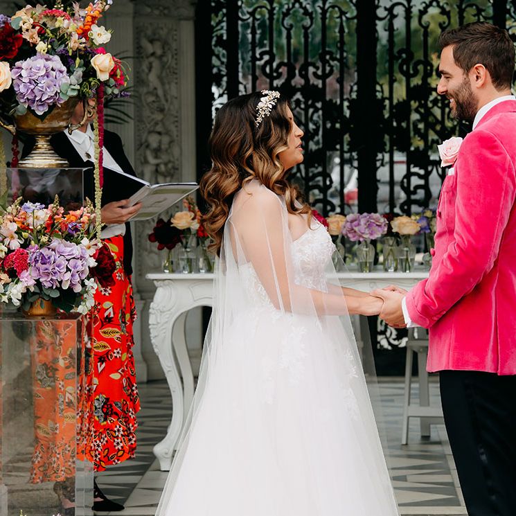 Groom in a vibrant pink velvet suit jacket holding hands with the bride in a strapless lace tulle wedding dress and sheer bridal cape for their civil ceremony 