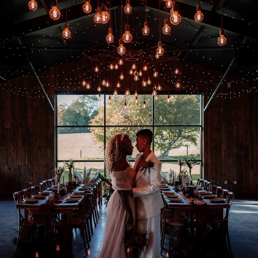 Bride in white silk suit and deep burgundy velvet crop top with autumnal dried floral boutonniere embracing bride in gold, star flecked embroidered dress and golden bridal crown in reception room at Silchester Farm with fairy lights and festoon chandelier lighting