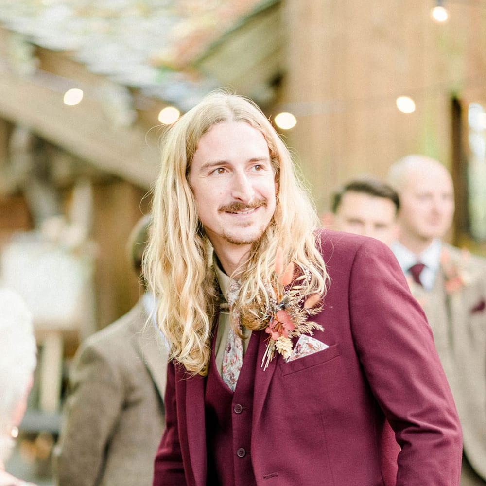 Groom wears maroon suit finished with floral pocket square and matching tie and dried floral buttonhole