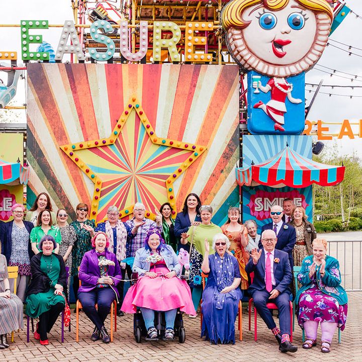Theme park wedding with brides in complimenting pink and purple bridal outfits sitting with wedding guests outside of a large colourful rollercoaster ride at Margate Dreamland 