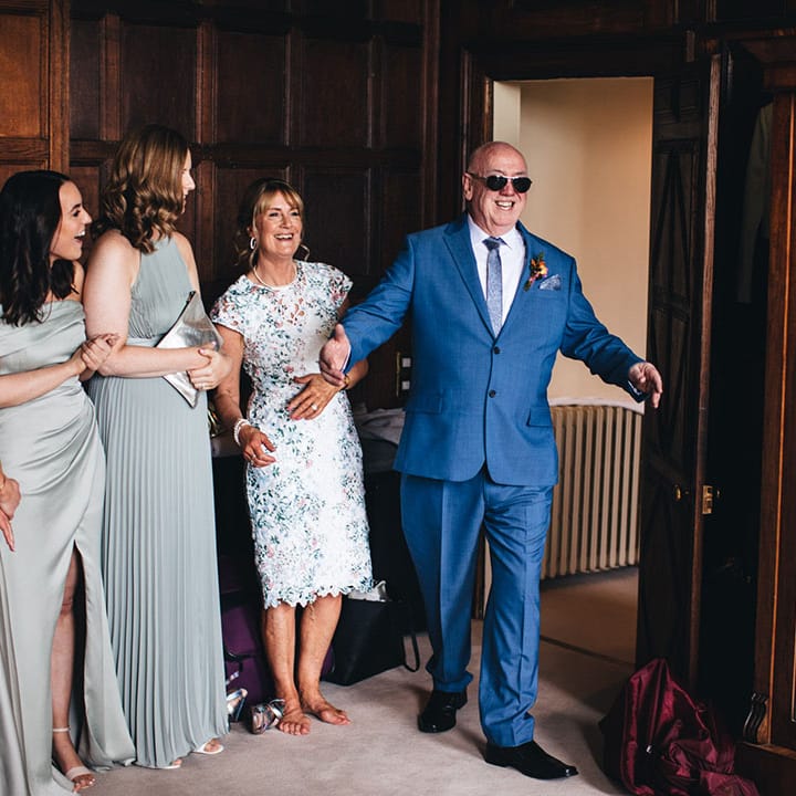 Father of the bride in a blue suit and pair of sunglasses walks into the room to get the first look at the bride 