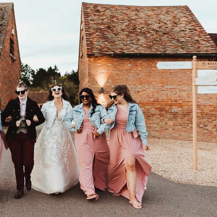 Bridal party wearing personalised denim jackets on top of dusky pink one shoulder bridesmaid dresses, illusion sleeve wedding dress and maroon bridal suit 