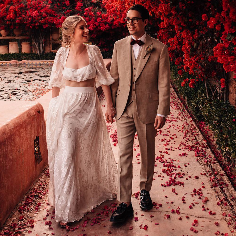 Bride in long sleeve crop top and lace maxi skirt walking hand in hand with the groom in three piece cream suit 