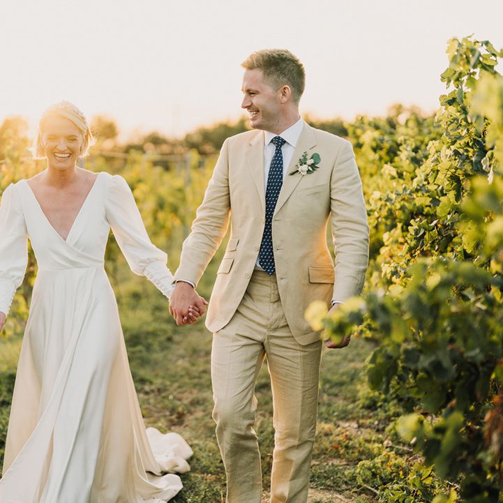 groom in a beige wedding suit holding hands with his bride in a floaty destination wedding dress