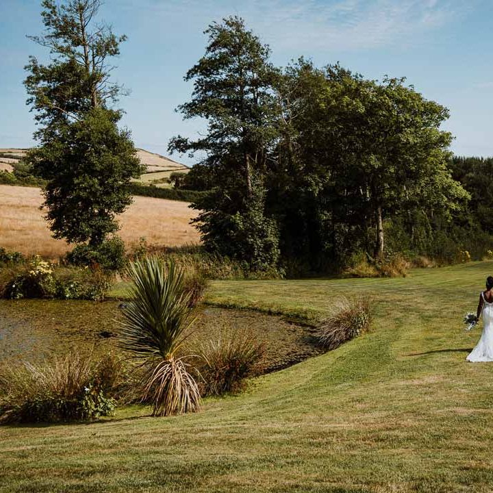 Bride in lace and mesh v back wedding dress with silver bridal hair accessories holding large white rose and foliage bridal bouquet walking hand in hand with groom in charcoal grooms suit on the grounds of Tredudwell Manor