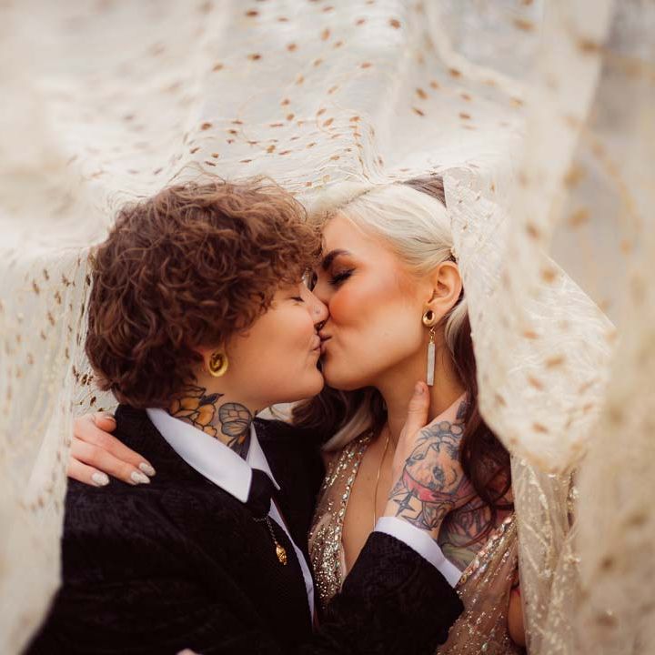 Bride and bride kissing underneath gold and sheer polka dot bridal veil at Kingscote Barn Tetbury LGBTQIA+ wedding 