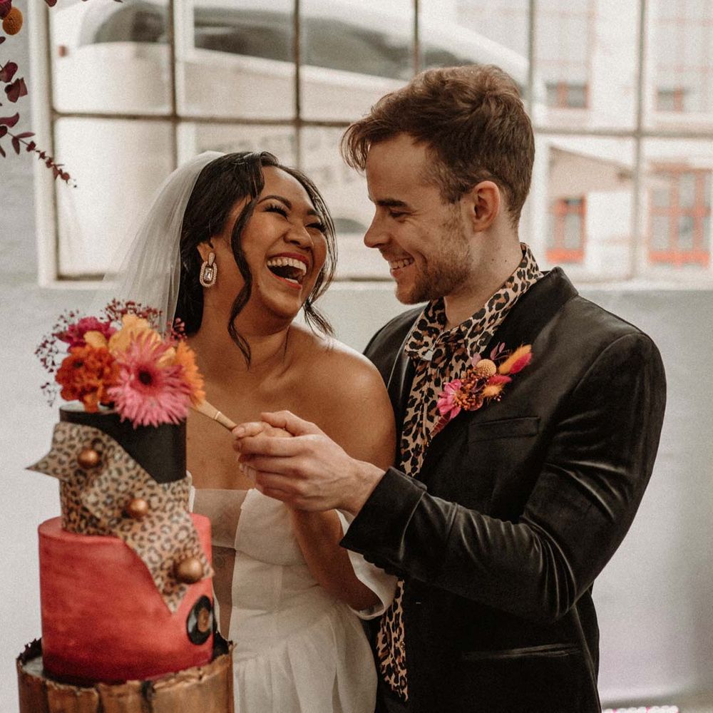 Bride in off shoulder tulle short wedding dress with church length veil and groom in wet look black grooms suit with leopard print shirt and pink and orange boutonniere cutting three tier alternative retro wedding cake with leopard print and flower toppers 