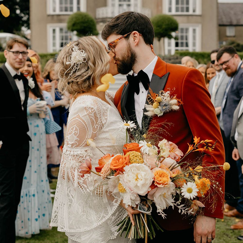 Groom in velvet suit jacket with bride in lace wedding dress carrying orange and yellow wedding bouquet 