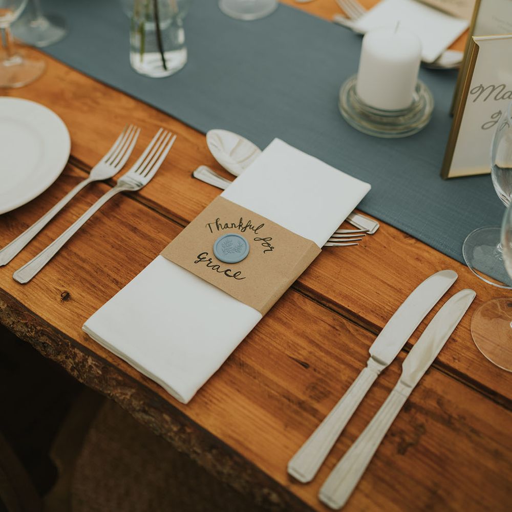 Blue wedding table runner on wooden table with white plates, white napkins with thank you sign with blue wax seal 