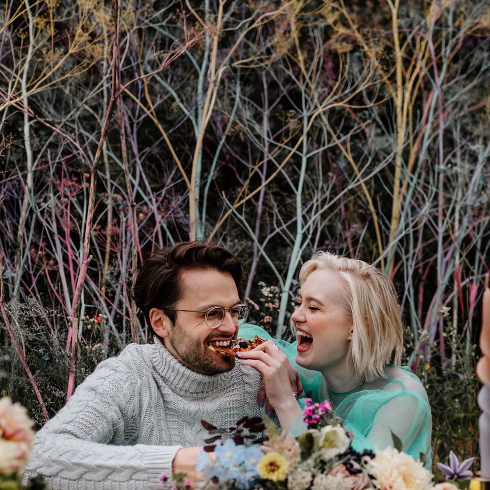 White blonde bride with bob length hair feeding her bearded groom in glasses pizza on the beach 
