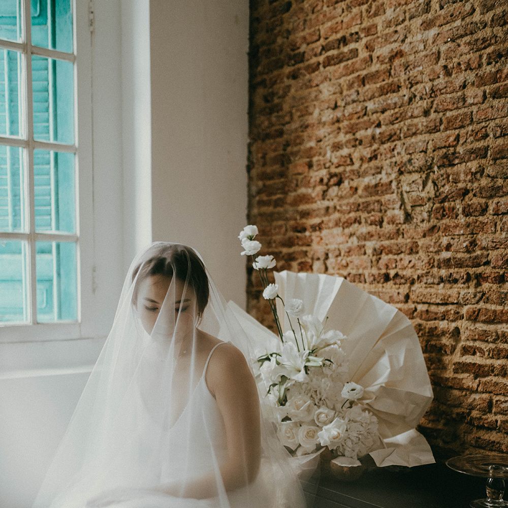 Asian bride in a fitted slip wedding dress sitting on a stool with a wedding veil over her face