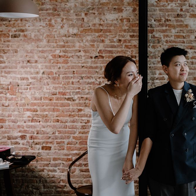 Bride in a fitted slip dress laughing holding hands with her bride in a double breasted navy suit jacket