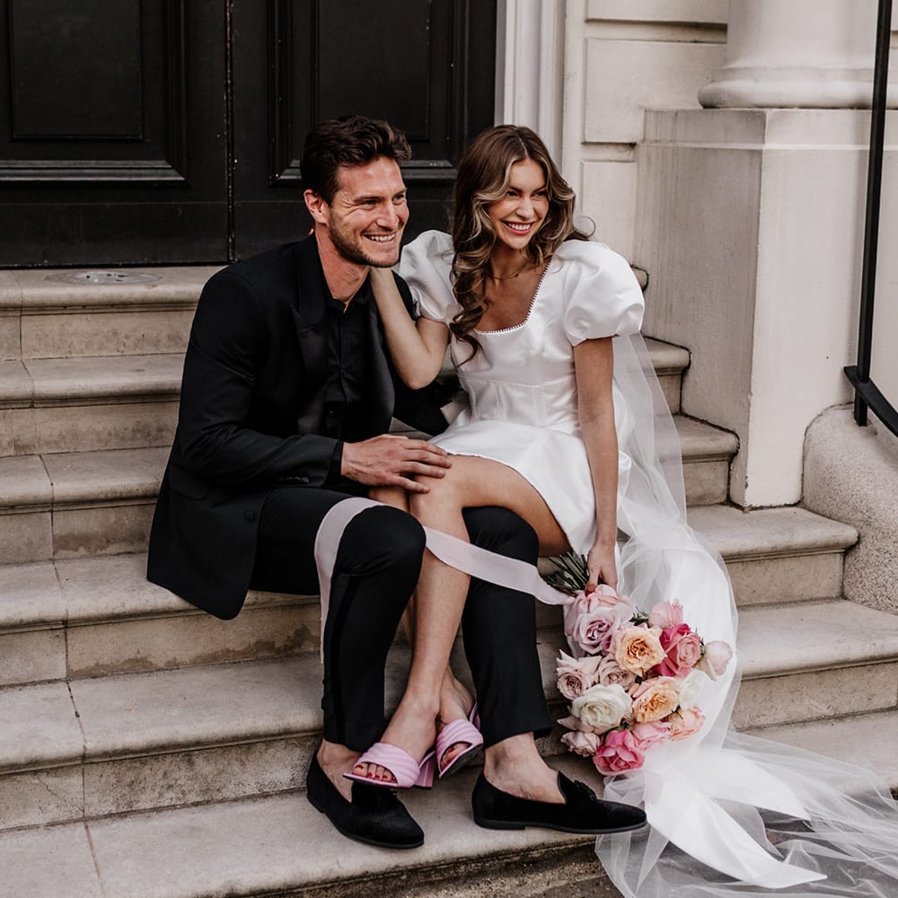 Stylish bride and groom sitting on some steps in Mayfair with bride in a short dress and pink shoes 