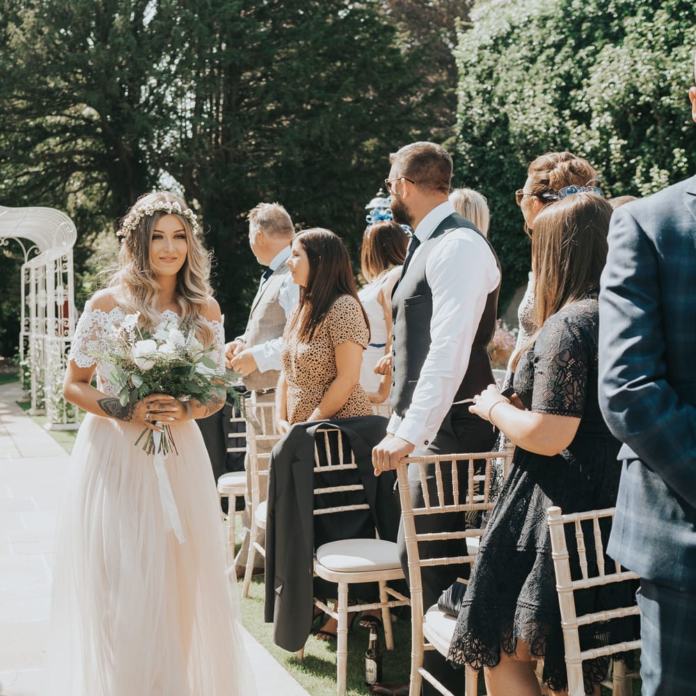 A bridesmaid with a bardot neckline dress holds a bouquet as she walks down an outdoor wedding aisle.