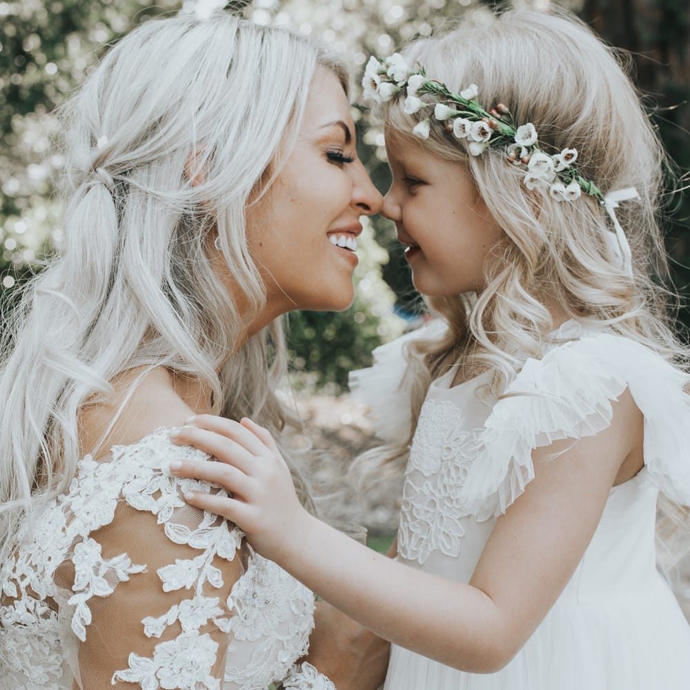 A bride is nose to nose with her daughter for a wedding portrait. They both have blonde hair. The little girl wears a flower crown.