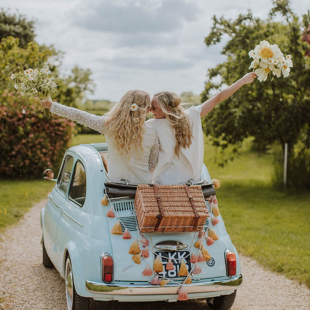 two brides riding a convertible fiat 500 wedding car with wicker basket and tassel decor on the back