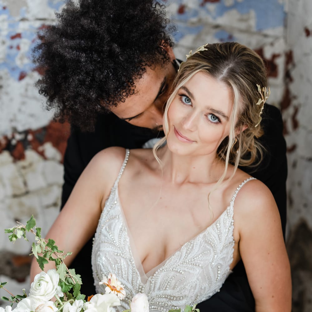 Bride with natural makeup in a beaded wedding dress with thin straps laying on her husbands lap 