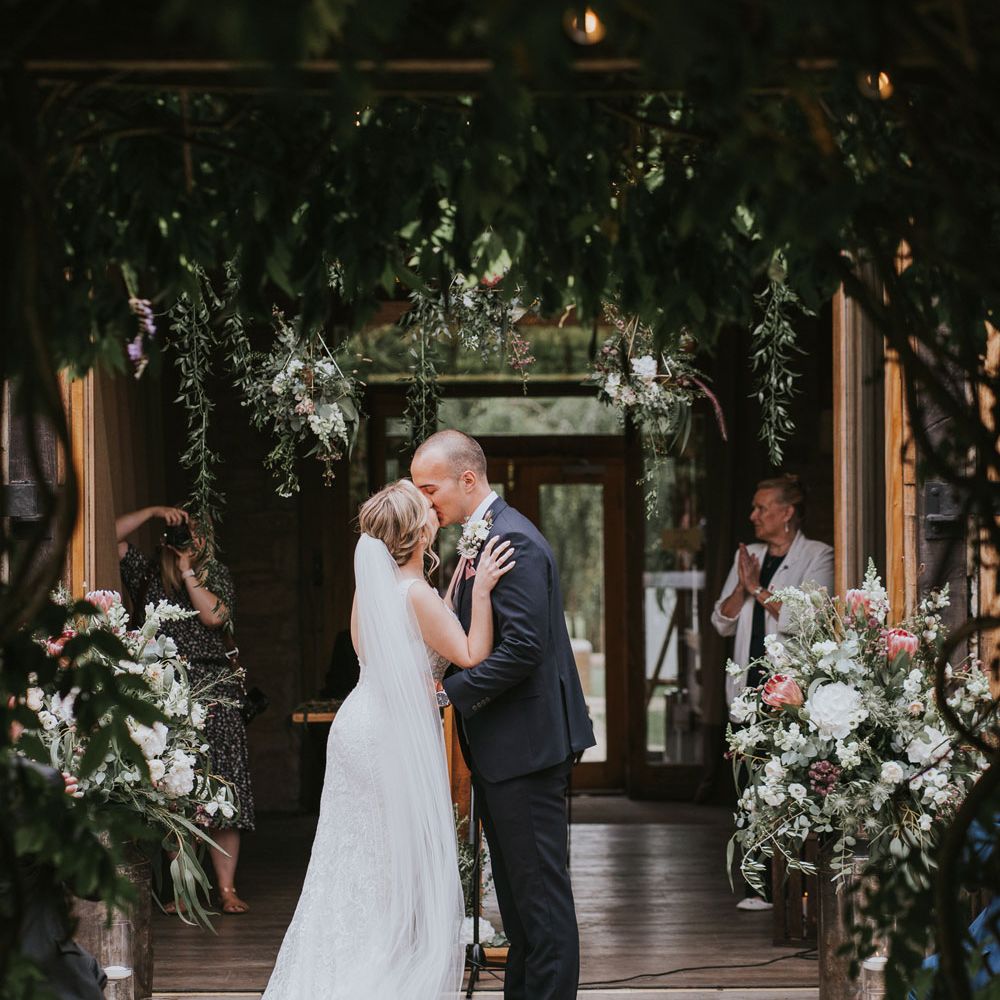 Bride in lace Justin Alexander wedding dress with train and veil kisses groom in dark three piece suit with buttonhole under hanging flowers during outdoor wedding ceremony at Tythe Barn